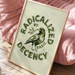 Op de afbeelding: Een ingelijste kunstdruk met een lichtgroene achtergrond. De print toont een groene vogel die een takje in zijn snavel houdt, omringd door de woorden "RADICALIZED BY BASIC DECENCY" in een cirkelvormig ontwerp. De lijst is licht van kleur.