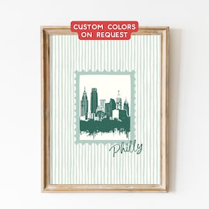 Könnte beinhalten: Gerahmter Kunstdruck mit einer grün-weißen Illustration der Skyline von Philadelphia in einem Briefmarkendesign. Das Wort "Philly" steht unter der Skyline. Der Hintergrund hat vertikale Streifen. Der Rahmen ist aus Holz.