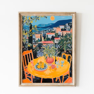May include: A framed art print depicting a sunny outdoor scene. An orange table is set with fruit, wine glasses, and a vase of greenery. Two orange chairs flank the table. The background shows a coastal town under a blue sky.