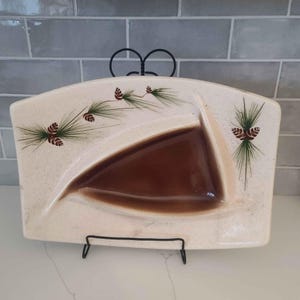 May include: A white ceramic serving dish with a brown triangular section and a pine tree design with brown pine cones. The dish is on a black metal stand.