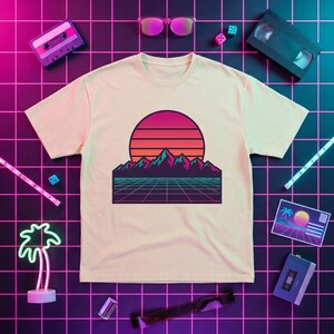 May include: A cream-colored t-shirt featuring a graphic print of a stylized mountain range under a large, striped sunset circle. The design includes a dark purple and pink color palette with a grid pattern at the base, evoking a retro aesthetic.