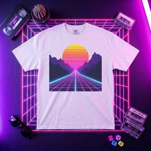 Peut inclure: T-shirt blanc avec un motif de coucher de soleil rétro, avec une route menant au soleil. Le graphisme utilise des tons de violet, rose, orange et bleu. Le t-shirt est entouré de cassettes et d'une cassette VHS, évoquant une esthétique rétro.
