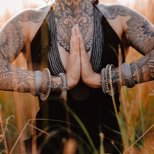 Könnte beinhalten: Eine Person mit komplizierten Tattoos an Armen und Brust sitzt in einem Feld mit hohem Gras. Sie trägt ein schwarzes Oberteil und silberne Armbänder. Ihre Hände sind in einer Gebetsposition zusammengelegt.