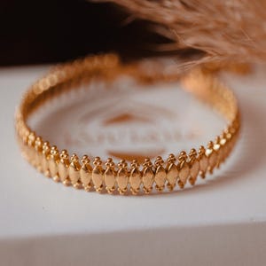 Peut inclure: Un bracelet doré avec un motif complexe de formes ovales répétées et de petites perles. Le bracelet est présenté sur une boîte blanche, avec un arrière-plan flou de plantes séchées.