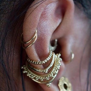 Puede incluir: Primer plano de una oreja adornada con múltiples pendientes dorados. La oreja presenta aros apilados, bandas con cuentas y un grupo decorativo de joyas doradas. También es visible un pequeño pendiente de disco adornado.