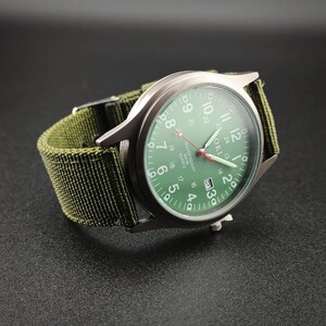 May include: A military-style wristwatch with a green face and a matching green nylon strap. The watch has a silver-colored case and features white numerals and hands. The watch face displays the brand name "OKI" and the words "WATER RESIST" and "QUARTZ".