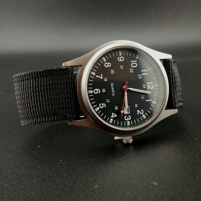 May include: A black analog watch with a silver-colored case and a black nylon strap. The watch face is black with white numerals and hands, and a red second hand. The watch is a quartz movement.
