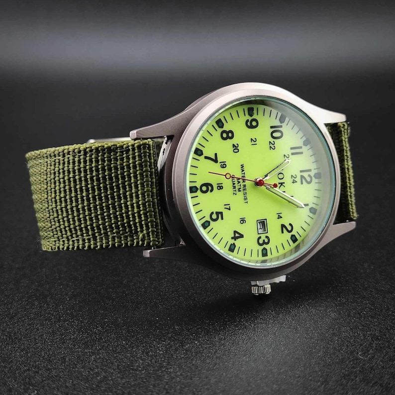 May include: A silver-toned wristwatch with a green face and a green fabric band. The watch face displays black numerals and hands, with the text "WATER RESIST ATM QUARTZ". The watch has a date display at the 3 o'clock position.