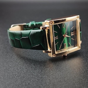 May include: A gold-toned rectangular wristwatch with a green face and a green leather band. The watch face features Roman numerals and gold hands. The band has a textured, crocodile-like pattern.