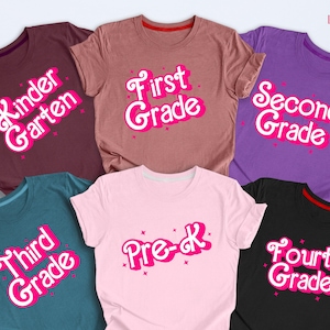 May include: A collection of colorful t-shirts, each displaying a grade level from Pre-K to Fourth Grade. The text is in a playful font with a pink outline and star accents. The shirts are in various colors, including burgundy, pink, purple, teal, and black.