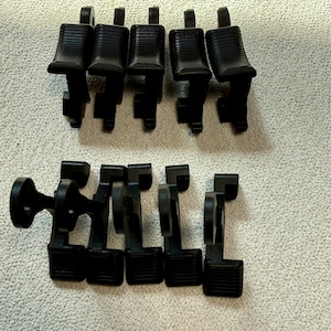 May include: A collection of black plastic furniture feet, arranged in two groups. The feet have a rectangular base with a ridged surface and a stem. One foot has a circular base. The items are on a light-colored, textured surface.