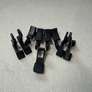 May include: A collection of black plastic clips, likely for a car or other mechanical device, are arranged on a light-colored surface. The clips have various shapes, including rectangular and curved designs, with textured surfaces.