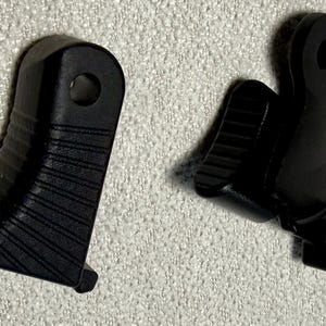 May include: Two black plastic components with a ridged design. Each piece has a hole and a small rectangular extension. The items are likely parts of a larger mechanism or device.
