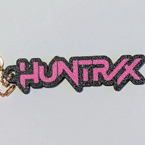 May include: A pink and black keychain with the word "HUNTRX" in a stylized font. The letters are pink with a glittery texture, outlined in black. A gold-coloured clasp and chain are attached to the keychain.