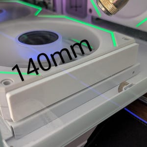 May include: A close-up of a white computer fan with green LED accents. The fan is labeled with "140mm". The fan is part of a computer case, with a clear side panel.