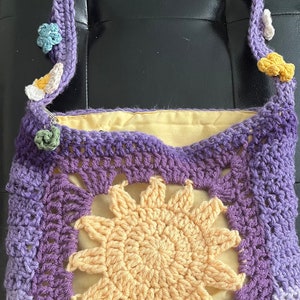 May include: A crocheted purple and yellow shoulder bag with a flower design. The bag has a yellow sun in the center and a purple and white flower design on the strap.