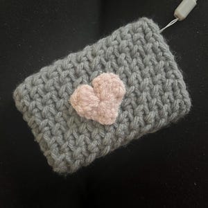 May include: A grey knitted pouch with a pink heart sewn on the front. The pouch has a grey cord attached to it.