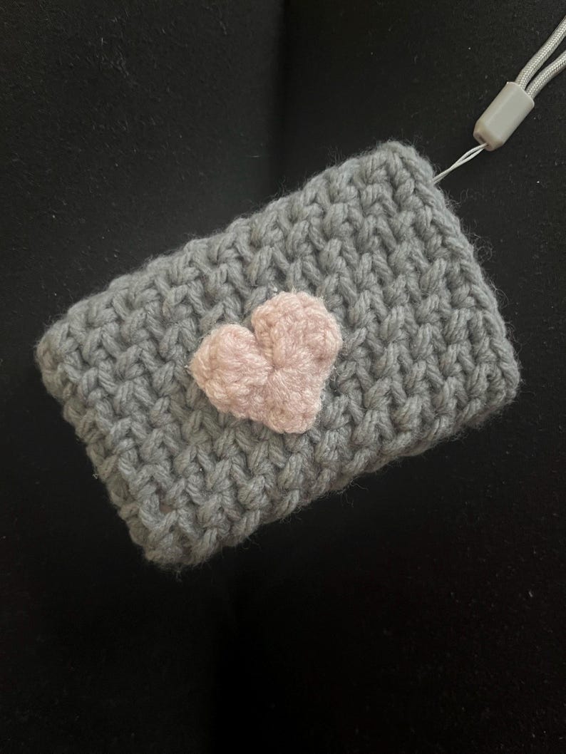 May include: A grey knitted pouch with a pink heart sewn on the front. The pouch has a grey cord attached to it.