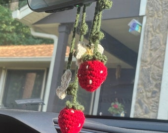 May include: Crocheted car charm featuring two red strawberries with white seed details, green stems, and cream-colored leaves. The charm hangs from a car's rearview mirror, adding a touch of handmade charm to the vehicle's interior.