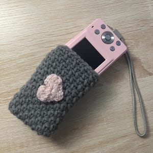 May include: A gray crocheted camera case with a pink heart design. The case is holding a pink digital camera with a screen and buttons.