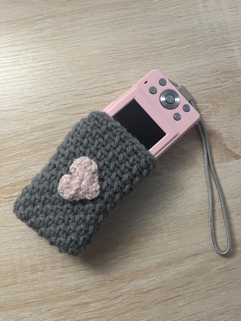 May include: A grey crocheted camera case with a pink heart design. The case is holding a pink digital camera with a screen and buttons.