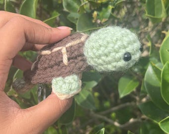 May include: A handmade crochet turtle in shades of brown and green. The turtle has a light green head and feet, a brown shell with tan accents, and small black eyes. The turtle is held in front of a background of green leaves.