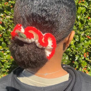 May include: A red, white, and green crocheted hair scrunchie is worn in a bun. The scrunchie has a wavy design with a red border and white and green accents. The person is wearing a black hoodie.