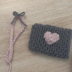 May include: A grey knitted cup cosy with a pink heart design and a pink and grey crocheted strap with a bow.