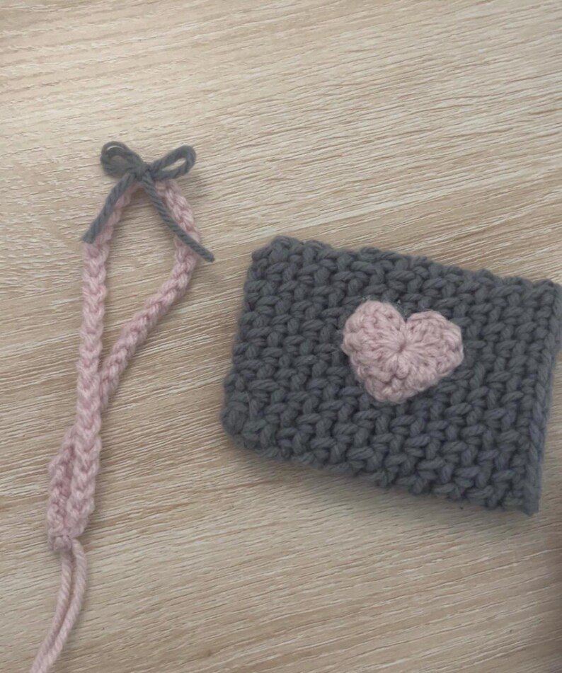 May include: A grey knitted cup cosy with a pink heart design and a pink and grey crocheted strap with a bow.