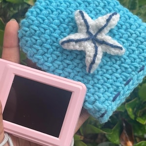 May include: A light blue crocheted case with a white and navy blue starfish applique. A pink digital camera with a silver strap is held in front of the case. The case is square and has a textured appearance.