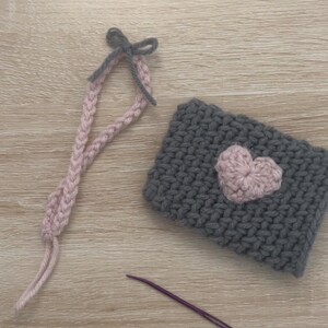 May include: A grey crocheted pouch with a pink heart design. The pouch has a grey and pink crocheted strap with a bow at the top. A purple crochet hook is also pictured.