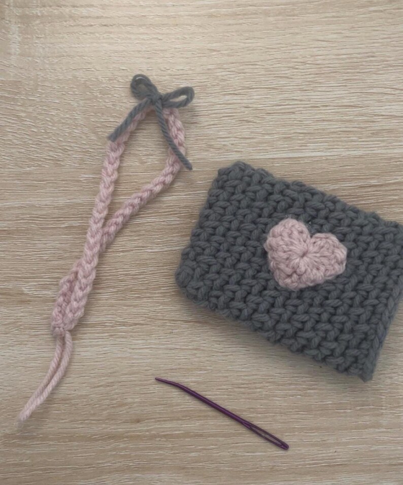 May include: A grey crocheted pouch with a pink heart design. The pouch has a grey and pink crocheted strap with a bow at the top. A purple crochet hook is also pictured.