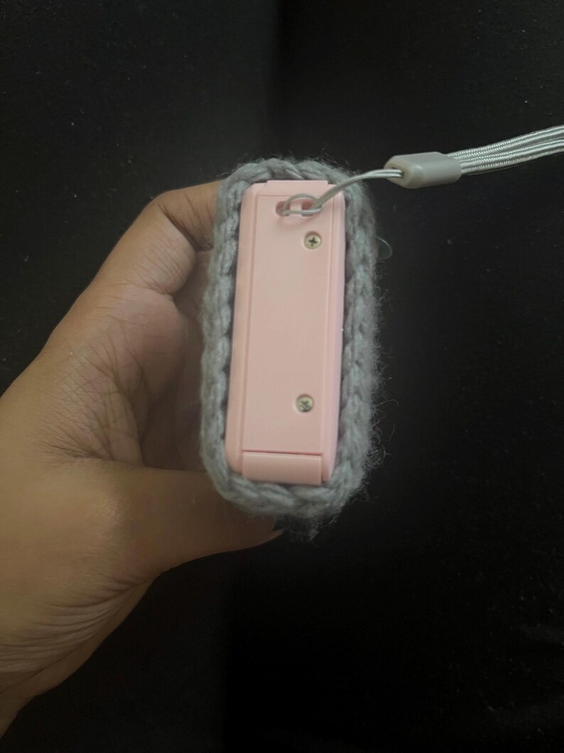 May include: A pink camera case with a grey crocheted cover and a grey strap.