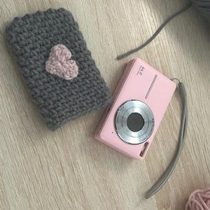 May include: A grey crocheted pouch with a pink heart design, a pink digital camera with a silver lens, and pink yarn.