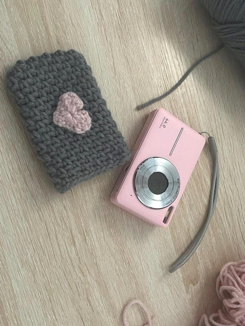 May include: A grey crocheted pouch with a pink heart design, a pink digital camera with a silver lens, and pink yarn.