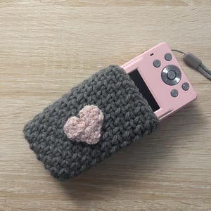 May include: A grey knitted camera case with a pink heart on the front. The case is covering a pink digital camera.