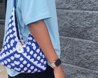 May include: A blue and white crocheted shoulder bag with a white button. The bag is worn by a person wearing a light blue t-shirt and black pants.