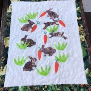 May include: A quilted wall hanging featuring a whimsical design of brown bunnies, orange carrots, and green grass on a white background. The quilt is bordered with a floral fabric and has a textured, quilted surface.