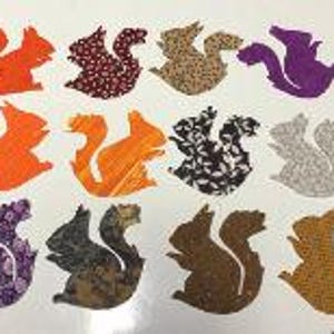 May include: A collection of twelve fabric squirrel cutouts arranged in three rows. The squirrels feature various patterns and colors, including solid orange, floral prints, polka dots, leaf motifs, and textured brown, purple, and gray fabrics.