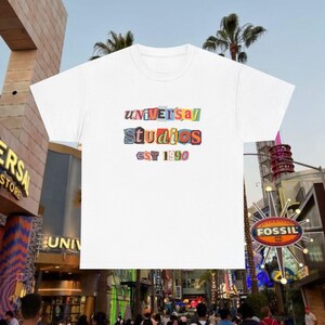 Puede incluir: Camiseta blanca con un estampado de colores "Universal Studios est 1990" en la parte delantera.