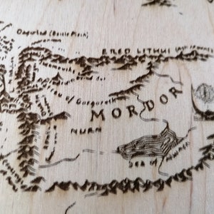 May include: A close-up of a wooden map of Middle-earth, showing the land of Mordor. The map is burned into the wood, with the text "Mordor" clearly visible. The text "Sea of Nurn" is also visible.