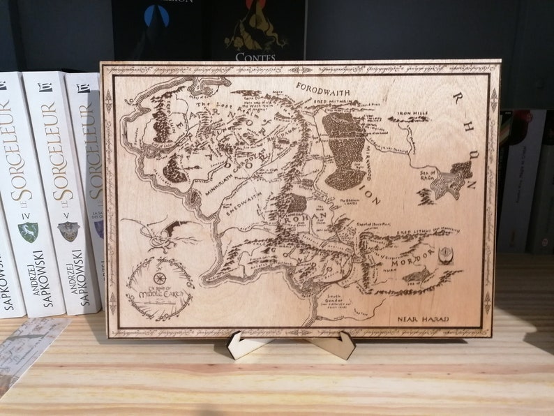 May include: A wooden map of Middle-earth from The Lord of the Rings, featuring the inscription "The Shire" and the text "The Lord of the Rings" in a circle. The map is mounted on a wooden stand.