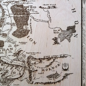 May include: A detailed map of Middle-earth from J.R.R. Tolkien's The Lord of the Rings, featuring the land of Mordor, the Iron Hills, and the Sea of Rh&ucirc;n. The map is printed on a light brown wooden surface.