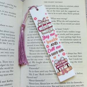 May include: A white bookmark featuring illustrations of books and bows in pink and brown, with the text "Buy me books AND Call me A GOOD GIRL". A pink tassel is attached. The bookmark is placed on the open pages of a book.