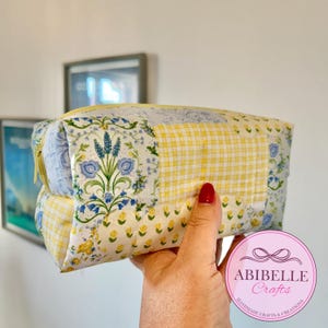 May include: A patchwork cosmetic bag with floral and gingham patterns in yellow, blue, and white. The bag has a yellow zip and is held in a hand. The bag is labelled "ABIBELLE Crafts Handmade Crafts & Creations."