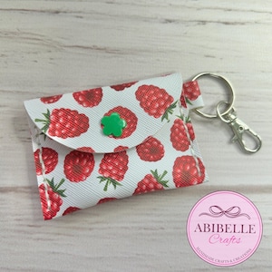 May include: A small, white wallet featuring a red raspberry pattern and a green flower-shaped button. It has a silver key ring and clasp. The design includes the text "ABIBELLE Crafts".