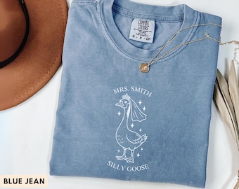 May include: A blue jean colored t-shirt with the words "MRS. SMITH" above a cartoon goose wearing a veil and crown. The words "SILLY GOOSE" are below the goose. A gold necklace is draped across the shirt.