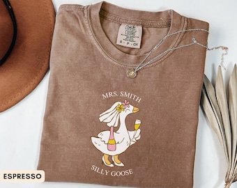 May include: Espresso-colored t-shirt with a graphic of a goose wearing a veil and holding a champagne glass and bottle. The text "MRS. SMITH" and "SILLY GOOSE" are printed on the shirt. A silver necklace is draped across the shirt.