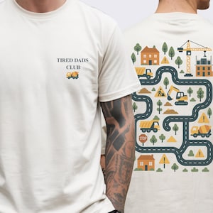 May include: Cream-colored t-shirt with the text "TIRED DADS CLUB" and a small truck graphic on the front. The back features a colorful construction-themed design with trucks, houses, trees, and a crane.