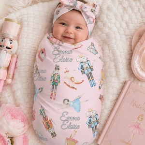May include: A baby swaddled in a light pink blanket featuring a Nutcracker-themed print with the name Emma Eloise, wearing a matching headband. The swaddle displays illustrations of nutcrackers, ballerinas, and mice, alongside the name Emma Eloise.